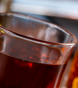 A macro close-up shot of a premium hotel drinkware glass cup with a smooth, thick rim, filled with dark amber liquid showing high-clarity glass details.