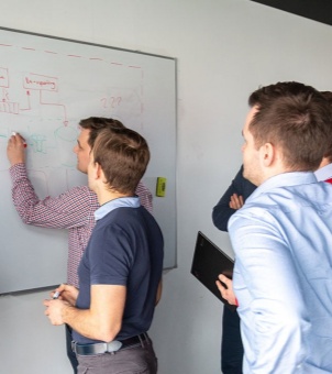 xinghuo-design-team-whiteboard-session A group of male engineers and designers collaborating during a whiteboard session. One team member draws a system architecture workflow with red and green markers, while others look on attentively, holding a tablet and analyzing the diagram.