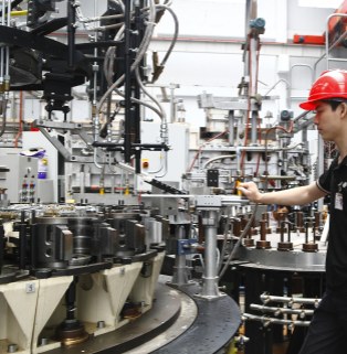 This is a close-up, medium-range realistic photograph taken inside a modern industrial plant, showcasing Xinghuo Glass's automated manufacturing capabilities and the details of its glass production line. On the left and center of the image stands a massive, complex, modern, multi-station rotary glass pressing or blowing machine. The steel frame is supported by numerous numbered, off-white conical mold bases, while above it are a dense network of metal pneumatic and hydraulic transmission pipes, mechanical rocker arms, spring suspension components, and a complex web of black control cables. The entire large machine exudes a robust industrial metallic feel. In the background, the spacious, tall white steel structure of the factory building and other large industrial furnaces are faintly visible. On the right side of the image, a young, capable Asian male technician stands sideways beside the machine, wearing a bright red standard construction helmet and black short-sleeved work clothes. His right hand rests on the silver metal control handle in the center of the machine, his expression focused and his gaze intently fixed on the operation of the automated turntable. The entire photo is realistically lit and without any artificial embellishment, perfectly integrating the cutting-edge, modern heavy-duty manufacturing equipment for everyday glass with rigorous and safe front-line production operations, fully demonstrating the strong foundation of this large foreign trade factory in terms of mass production and fulfilling large-volume orders.