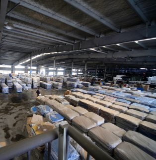 This is a hardcore, realistic photograph taken from a high-angle, overhead perspective, designed to showcase the massive industrial warehousing and reliable wholesale supply chain of a major B2B export manufacturer. The main subject of the image is an extremely spacious, tall, and seemingly bottomless modern steel-structured logistics warehouse hall. The high ceiling is supported by dense gray I-beams and metal trusses, with rows of industrial spotlights hanging in the depths, projecting bright white ribbons of light. On the entire open warehouse floor, thousands upon thousands of massive quantities of goods are densely and meticulously stacked, arranged in dozens of long matrix lines: the goods in the foreground and midground on the right are tightly wrapped in thick, light brown moisture-proof kraft paper and transparent bubble wrap, stacked into rows of half-meter-high rectangular stacks; the goods in the background on the left are wrapped in blue and white striped rainproof high-strength plastic protective film, also arranged in rows and matrices extending endlessly towards the light-filled door at the far end of the warehouse. On the left, in a spacious, sturdy gray concrete walkway marked with signs of work and dampness, a warehouse worker in dark clothing moves almost microscopically between the cargo areas, emphasizing the sheer size of the warehouse space. A section of metal railing juts out at an angle in the foreground. The photograph, with its strong contrast of light and shadow, profound sense of depth, and impressive scale of goods, vividly portrays the efficient logistics of a modern large-scale factory, its massive inventory, and its robust multinational supply chain ensuring stable, high-volume delivery.