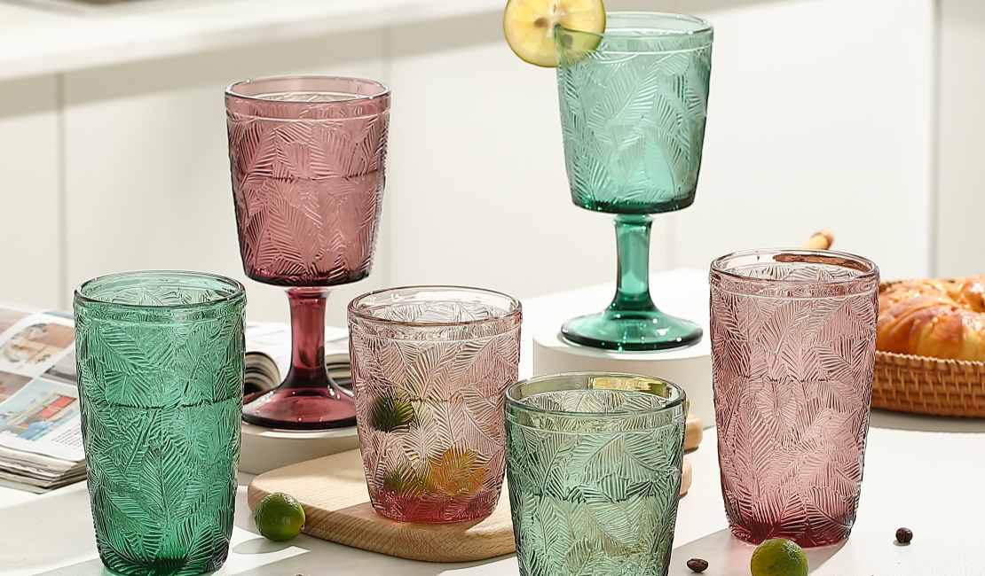 A stylish and eclectic collection of high borosilicate solid color glass tumblers from the Solid Color Glass Tumblers set.jpg collection is artfully arranged on a kitchen counter, showcasing intricate embossed palm leaf patterns. This six-piece set includes pairs of tall highball tumblers and shorter water glasses, as well as stemmed goblets in both purple-rose and teal-green hues. The glasses are displayed alongside kitchen staples like a cutting board, small limes, a magazine, and a basket of croissants, highlighted by soft natural light filtering through, emphasizing the texture and color of the handmade glassware for home decor.